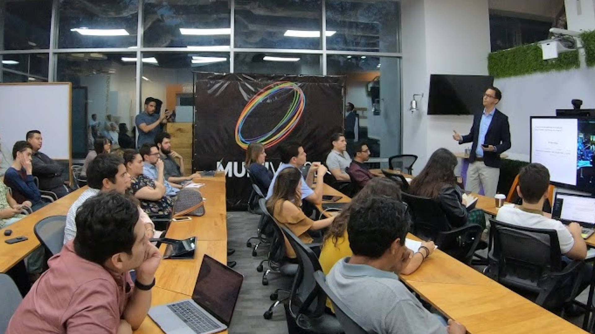César Rengifo leading a workshop in a bright room with participants seated at tables with laptops, facing a presentation screen.